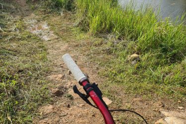A view from the handlebars of a mountain bike, overlooking a grassy path leading down to a serene pond surrounded by lush greenery. In the background, a small residential area is visible, with houses and trees under a partly cloudy sky. Bukit batu trail park mountain bike trail.