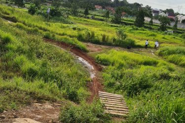 A scenic view of a grassy landscape with tall green grass and scattered trees. In the foreground, there is a dirt path with a wooden bridge crossing a small stream. In the background, power lines and telecommunications towers are visible, along with a few buildings. Two people can be seen walking in the distance. The sky is partly cloudy, suggesting a calm day. Bukit batu trail park mountain bike trail.