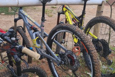 Two mountain bikes are parked beside a railing in a grassy area. The bike on the left is gray with yellow accents, while the bike on the right is bright yellow and black. Both bikes have muddy tires, indicating recent use on a trail. In the background, there are trees and a hint of a cloudy sky. Bukit batu trail park mountain bike trail.