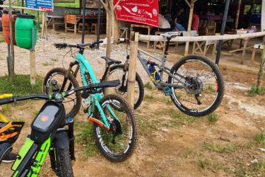 Three bicycles are parked in front of a small, rustic eatery with a thatched roof, signified by a blue and red banner that reads "Saung Bukit Batu Pak Eko." The scene is set in a natural environment, characterized by dirt paths and a few sparse trees. One bicycle is black and green, while the others are aqua and gray, juxtaposed against the earthy tones of the surroundings. Bukit batu trail park mountain bike trail.
