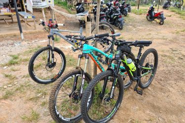 A pair of mountain bikes parked next to a tree, with a variety of motorcycles visible in the background. The scene is set in a grassy area with a dirt path, and there are signs indicating rules related to helmet use. The bikes are equipped for off-road riding, with prominent tires and accessories, including a water bottle. Bukit batu trail park mountain bike trail.