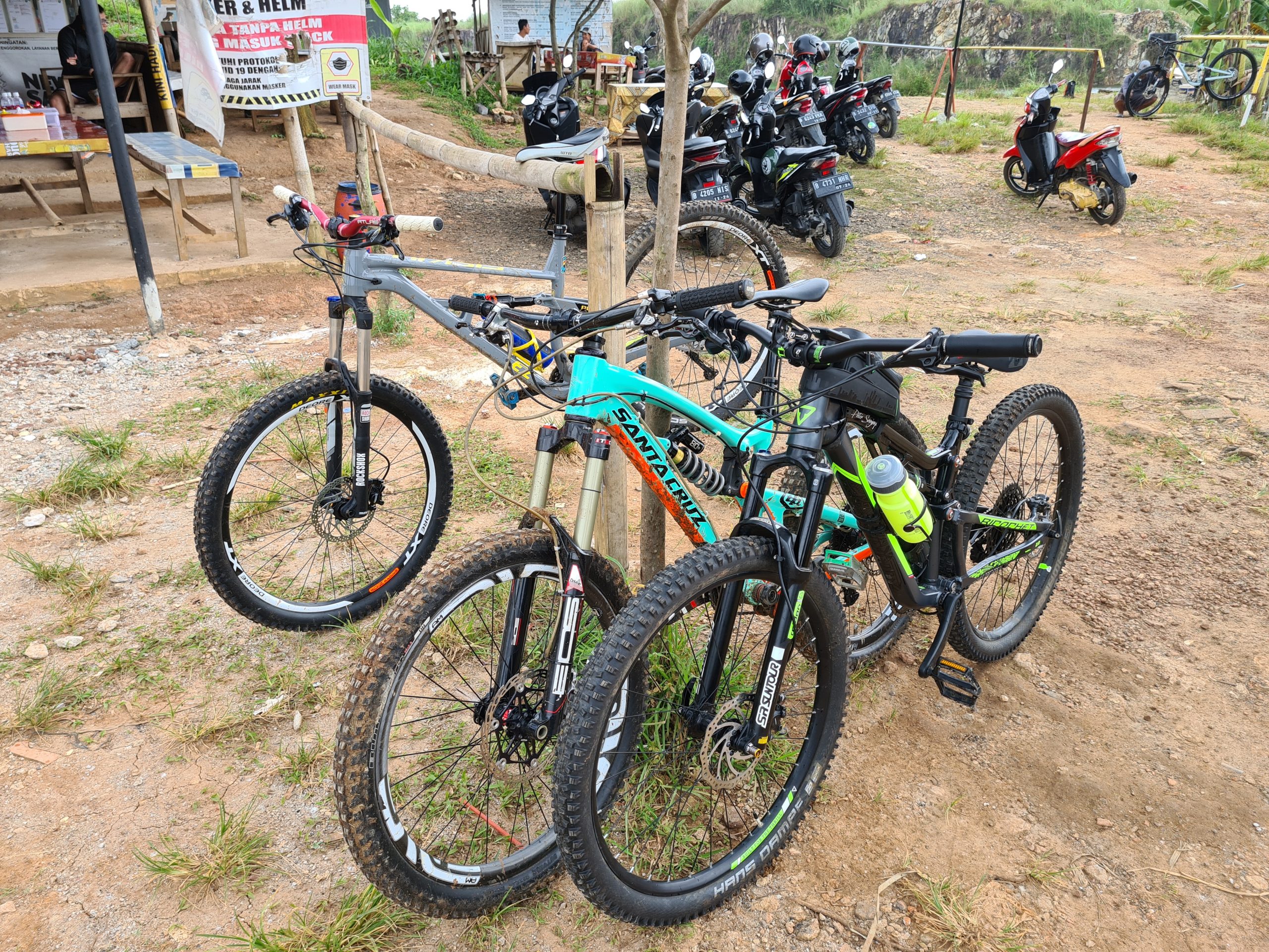 A pair of mountain bikes parked next to a tree, with a variety of motorcycles visible in the background. The scene is set in a grassy area with a dirt path, and there are signs indicating rules related to helmet use. The bikes are equipped for off-road riding, with prominent tires and accessories, including a water bottle. Bukit batu trail park mountain bike trail.