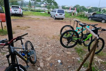 A view of a gravel parking area with bicycles parked on a wooden rack in the foreground. Two mountain bikes are visible, one with a gray frame and the other in bright green and black. In the background, several cars are parked, and there are trees and houses in the distance under a cloudy sky. Bukit batu trail park mountain bike trail.