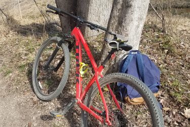 A red mountain bike leaning against a tree in a wooded area, with a blue backpack visible on the ground nearby. The ground is covered in leaves and twigs, and a small stream can be seen in the background. Rosaryville State Park mountain bike trail.
