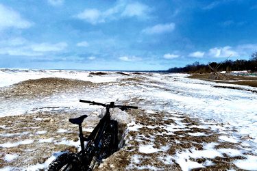 A black bicycle partially embedded in snow and ice on a winter landscape, with a clear blue sky and fluffy white clouds in the background. The scene shows a vast expanse of snow-covered terrain, emphasizing the bicycle's rugged tires designed for snowy conditions. The Beach mountain bike trail.