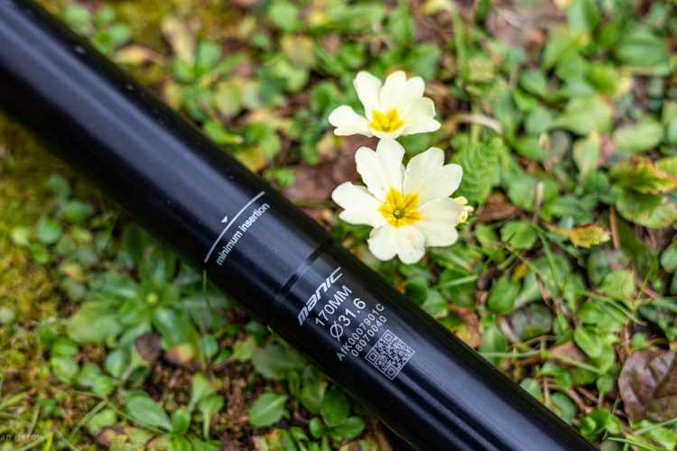 A black bicycle seat post with visible markings, including the text "minimum insertion" and "manic 110MM Ø 31.6," is resting on green grass along with small yellow and white flowers.
