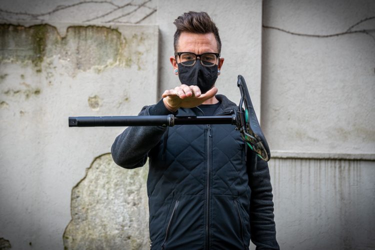 A person wearing a mask and glasses is performing a trick with a scooter, balancing it horizontally on one hand against a textured, outdoor background.