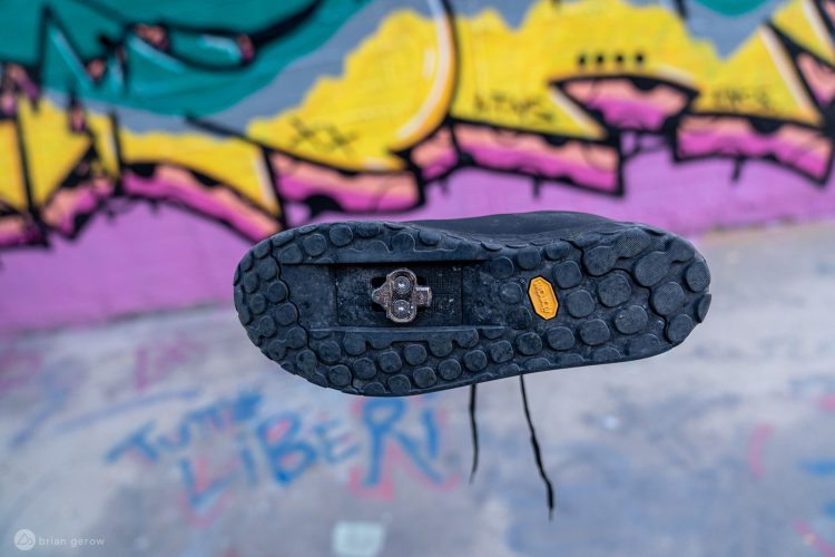 A close-up view of the sole of a black shoe, showcasing its textured design and a cleat attachment, with vibrant graffiti art in the background, including colorful shapes and the word "LIBERI."