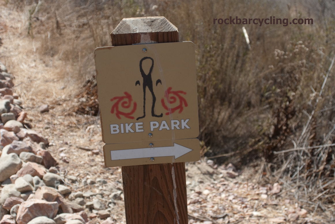A wooden sign indicating a bike park, featuring a stylized figure and swirling designs. The sign includes the words "BIKE PARK" and an arrow pointing to the right, with a backdrop of dry grass and rocks. Sapwi Trails Community Park to China Flat mountain bike trail.