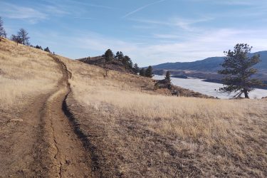 A winding dirt trail leads through dry grass and sparse trees, ascending a hillside with a view of a wide river and distant mountains under a clear blue sky. Reservoir Ridge mountain bike trail.