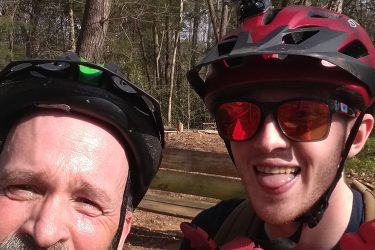 Two cyclists posing for a selfie in a wooded area. One person has a beard and is wearing a black helmet, while the other has a red helmet with sunglasses and a GoPro camera mounted on top. Both are smiling and making playful gestures, enjoying their time outdoors. San-lee Park mountain bike trail.