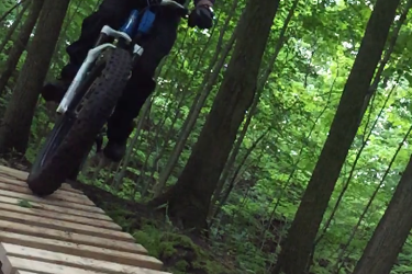 A mountain biker navigating a wooden plank trail through a green forest, wearing a helmet and black riding gear, with trees in the background. Swance Drain mountain bike trail.