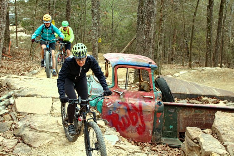 Three mountain bikers navigate a rocky trail in a wooded area, passing by a partially visible old, rusted truck. The lead biker, wearing a black jacket and helmet, is focused as they climb over rocks. The other two bikers follow behind on their bikes. The scene captures the adventurous spirit of mountain biking against a backdrop of trees and natural terrain.