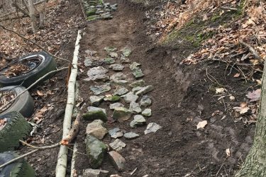 A dirt hiking path winding through a wooded area, with a trail made of stones and tire remnants on the side. Dried leaves cover the ground, and trees can be seen in the background, indicating a natural, outdoor setting. Swance Drain mountain bike trail.