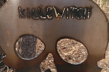 A large, metal skull-shaped sign with the word "KILLSWITCH" cut out at the top. The sign is mounted on a post in a wooded area, with trees in the background and dried leaves on the ground. There is also a small "Danger" sign visible nearby, adding to the ominous atmosphere of the location. San-lee Park mountain bike trail.