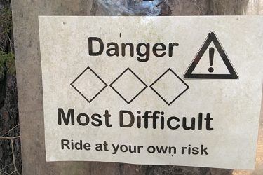 Sign attached to a tree indicating a difficult and potentially dangerous trail, with the text "Danger Most Difficult Ride at your own risk" and warning symbols. San-lee Park mountain bike trail.