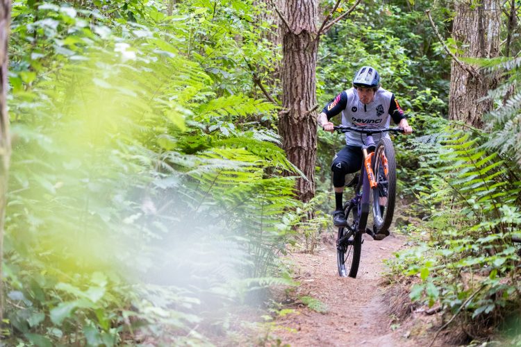 A mountain biker performing a wheelie on a trail surrounded by dense greenery and ferns, showcasing a dynamic action shot in a natural outdoor setting.