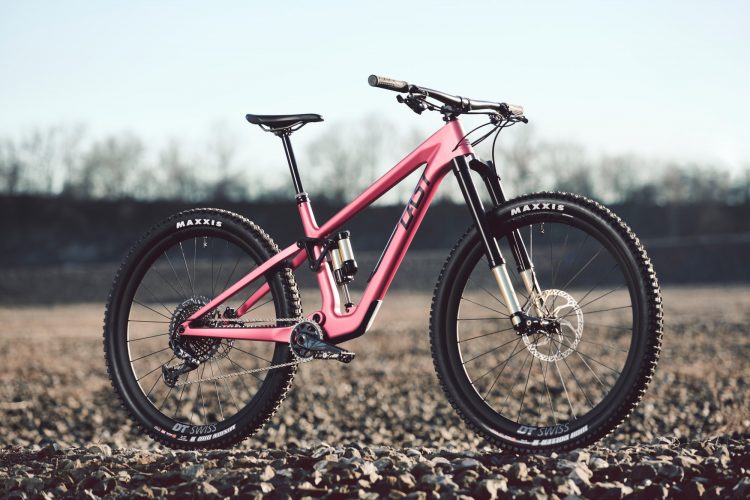 A pink mountain bike with a sleek design parked on a gravel surface, showcasing its sturdy tires and suspension system against a clear sky in the background.