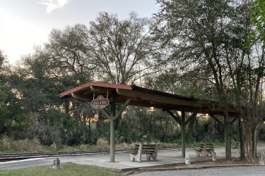 A small train station shelter with a red roof and a sign reading "DeLand." The area is surrounded by trees and vegetation, with two concrete benches under the shelter and a view of railroad tracks nearby. The sky shows early evening colors, suggesting a serene outdoor atmosphere. Lake Beresford Park Nature Trail mountain bike trail.