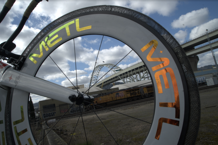 A close-up view of a bicycle wheel with the word "METL" prominently displayed in reflective letters. The background features a bridge and cloudy sky, along with elements of an industrial setting, including rail tracks and buildings.