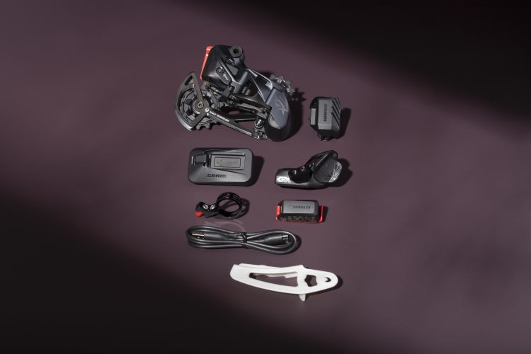 A collection of bicycle components arranged on a dark surface, including a derailleur, battery, wireless control unit, charging cable, and installation tool, all displaying the SRAM branding.