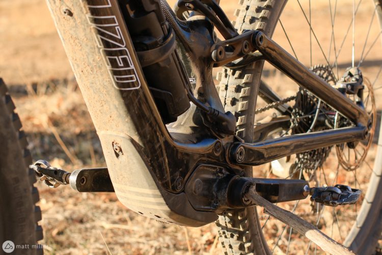 Close-up of a specialized mountain bike's drivetrain, showcasing the crankset, rear suspension linkage, and a slightly dirty frame against a blurred natural backdrop.