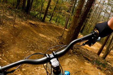 A close-up view of a mountain bike's handlebars on a dirt trail surrounded by trees, with a focus on the rider's gloved hand controlling the bike. The ground is covered with fallen leaves, and the trail curves ahead, suggesting a downhill path in a natural outdoor setting. Moontower mountain bike trail.
