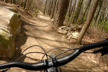 View from the handlebars of a mountain bike on a winding dirt trail through a wooded area, with large rocks and trees lining the path. The trail is surrounded by a mix of bare branches and budding greenery, suggesting early spring. Moontower mountain bike trail.