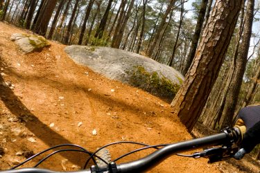 A view from a mountain bike navigating a dirt trail in a wooded area, featuring a large rock on the side of the path and trees in the background. The handlebars of the bike are visible in the foreground, indicating an action shot of downhill biking. Moontower mountain bike trail.