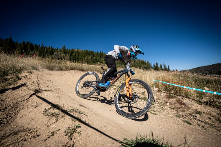 A mountain biker leans into a dirt track while performing a maneuver on a sunny day. The rider is wearing a blue helmet and gear, and the surrounding landscape features tall green trees and a clear blue sky. Dust is kicked up from the bike's tires as it navigates the terrain.