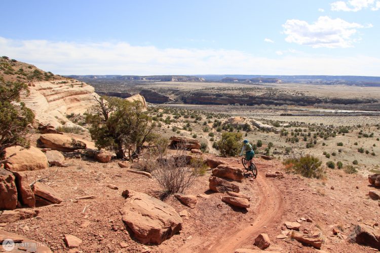 A cyclist navigates a winding dirt trail on a rocky terrain, surrounded by a vast landscape of cliffs and valleys under a clear blue sky with scattered clouds.