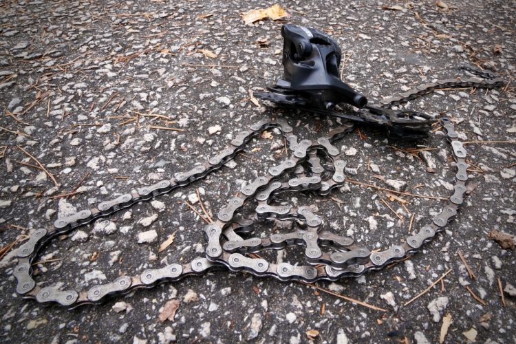 A close-up image of a bicycle chain lying on a gravel surface, partially tangled and connected to a black derailleur. Small twigs and leaves are visible around the chain, indicating an outdoor setting.