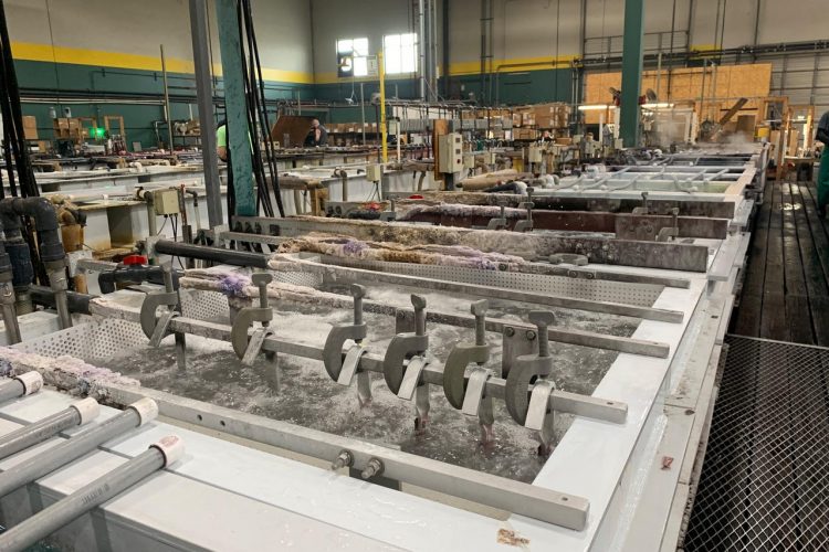 A large industrial workspace featuring multiple tanks filled with water, with mechanical apparatus and hoses. The area appears to be well-lit and organized, with various tools and equipment situated around the tanks. Workers can be seen in the background, engaged in activities related to the operation. The interior has high ceilings and walls painted in green and yellow, with stacked materials and boxes visible.