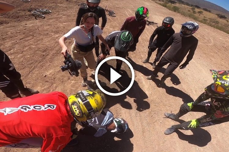 A group of motocross riders, wearing helmets and protective gear, gather around a fallen rider on a dirt track. A woman with a camera is capturing the moment. The scene takes place in an outdoor setting with hills in the background and several bicycles visible nearby.