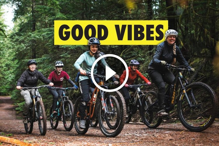 A group of five people riding mountain bikes on a forest trail, surrounded by tall, green trees. They are smiling and dressed in casual outdoor clothing and helmets. The text "GOOD VIBES" is displayed prominently at the top of the image, along with a play button icon in the center. The setting conveys a sense of adventure and camaraderie.