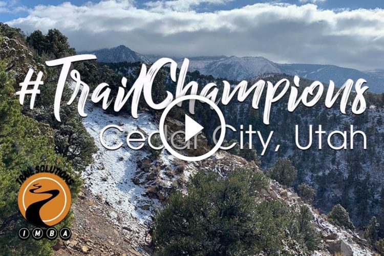 A scenic landscape of Cedar City, Utah, featuring a snowy mountain trail surrounded by lush greenery and distant mountains under a partly cloudy sky. The overlay includes the text “#TrailChampions” and a video play button. An emblem featuring a stylized trail logo is also visible.
