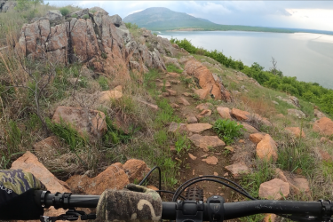 Photo of a mountain biker's perspective while navigating a rocky trail along a hillside. The foreground shows the handlebars of a mountain bike, with rough terrain and orange rocks visible. In the background, there is a lush green hillside leading down to a lake, under a partially cloudy sky. Lake Lawtonka Trails mountain bike trail.