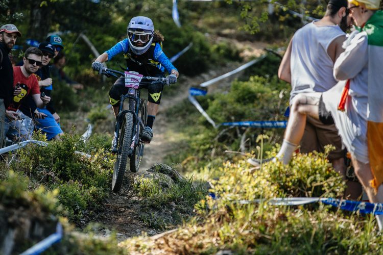 A mountain biker navigates a challenging trail surrounded by spectators. The rider, wearing a helmet and protective gear, is in mid-action, tackling uneven terrain with plants and rocks. In the background, cheering onlookers watch the event, with some holding drinks and enjoying the race atmosphere. Blue tape marks the course path in a forested setting.