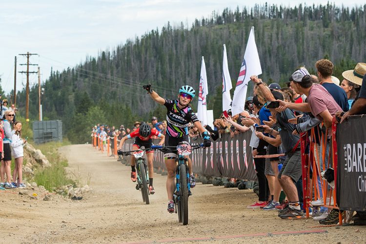 A mountain bike racer crosses the finish line, raising one arm in victory. A crowd of enthusiastic spectators cheers along the sidelines, showing expressions of excitement and support. The setting features a dirt path surrounded by trees and a clear sky, with banners and flags in the background.