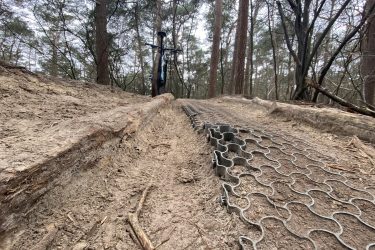 A view of a forest path with a mountain bike leaning against a tree. The trail shows a mixture of dirt and a grid-like matting laid along part of the path to provide traction. Surrounding trees are bare, indicating an early spring or late autumn setting, and the scene is somewhat overcast. Appelscha Rood-Blauw-Rood mountain bike trail.