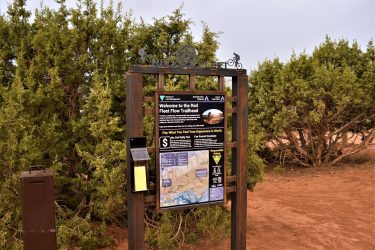 Sign at the Red Fleet Flow Trailhead, featuring information about the trail, fee payment options, and a map. Surrounded by green shrubs and under a cloudy sky. Red Fleet Downhill Flow Trail mountain bike trail.