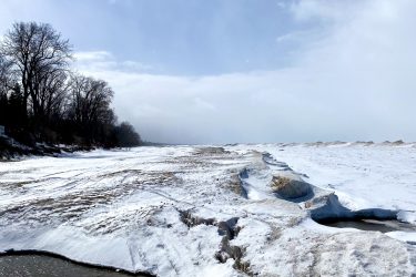 A snowy landscape featuring a frozen shoreline with patches of open water and snow-covered ground. In the background, trees line the edge of the shoreline under a partly cloudy sky. The Beach mountain bike trail.