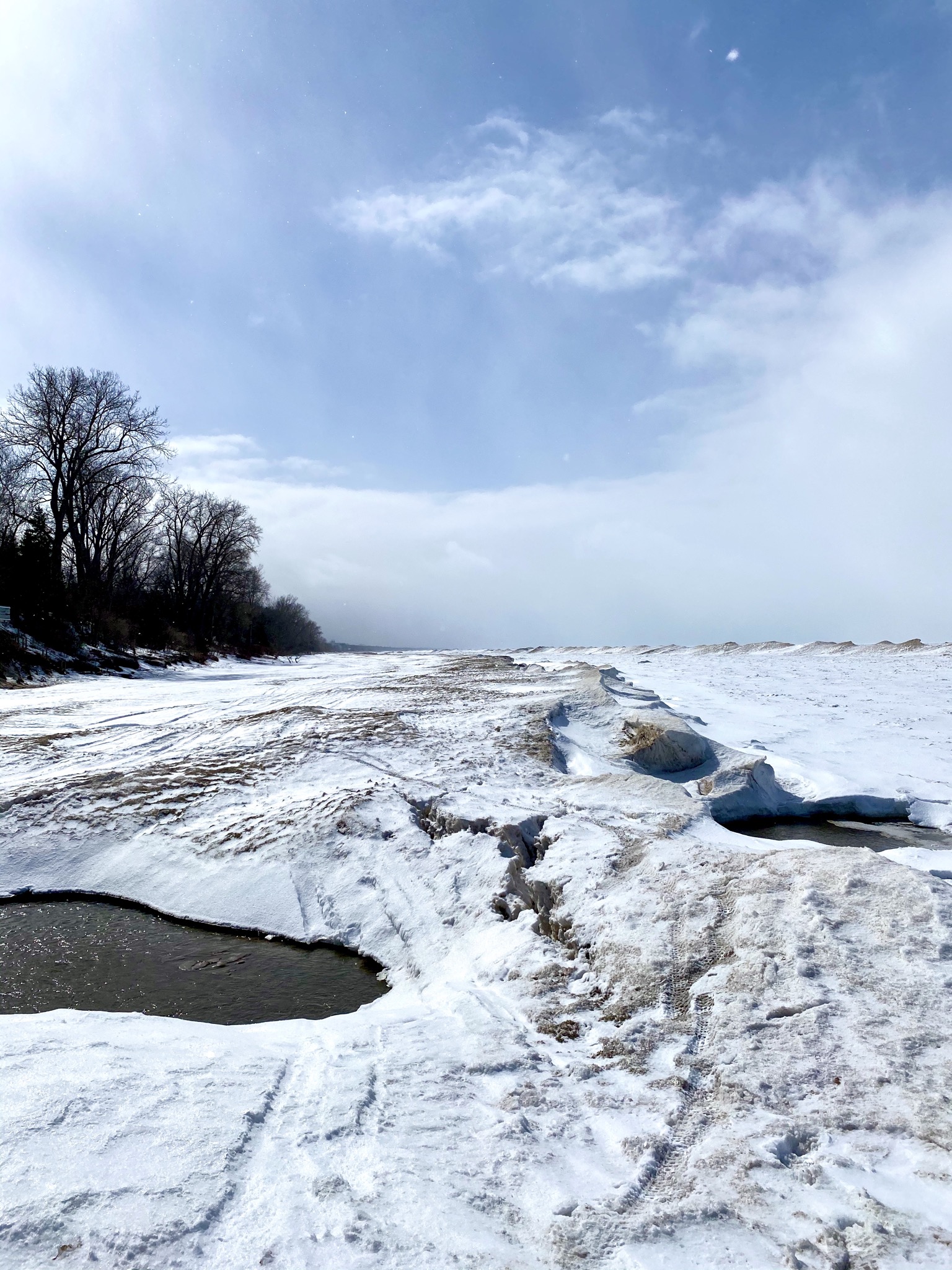 A snowy landscape featuring a frozen shoreline with patches of open water and snow-covered ground. In the background, trees line the edge of the shoreline under a partly cloudy sky. The Beach mountain bike trail.