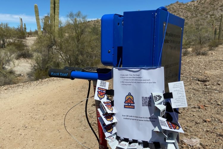 A blue trail sign mounted on a post in a desert setting, surrounded by cacti and mountain terrain. The sign features a QR code, instructions for trail users, and various small tags attached under the sign, likely providing information or promoting local services related to the trail. The sky is clear and blue, indicating a sunny day.