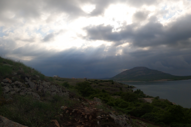 A scenic landscape featuring a rocky hillside with green grass, leading down to a calm lake. In the background, a mountain rises under a dramatic sky filled with dark clouds and rays of sunlight peeking through, creating a moody atmosphere. Small structures are visible in the distance along the shore. Lake Lawtonka Trails mountain bike trail.