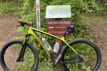A mountain bike positioned next to a trail sign indicating "Forest Lake," with a map of the trail and a "One-Way for Bikes" notice. The background features lush greenery typical of a forest setting. The bike is equipped with two water bottles and a small saddle bag. Bauxite Ridge mountain bike trail.