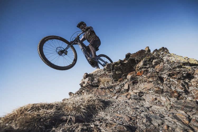 A mountain biker performing a wheelie on a rocky outcrop against a clear blue sky. The cyclist is dressed in black gear, showcasing dynamic movement and skill on the challenging terrain.