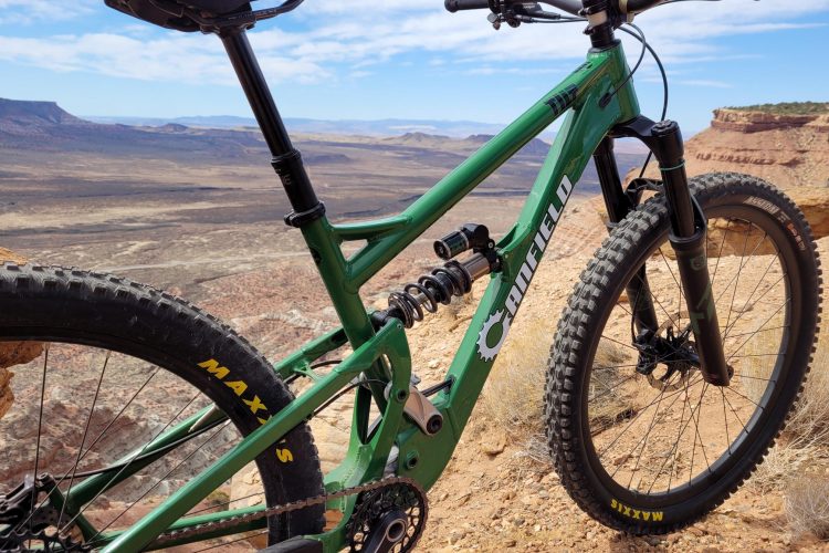 A close-up view of a green mountain bike with a matte finish, positioned on a rocky ledge overlooking a vast desert landscape. The bike features prominent Maxxis tires and a dual suspension system. In the background, faint mountain ranges and a clear blue sky complete the scenic view.