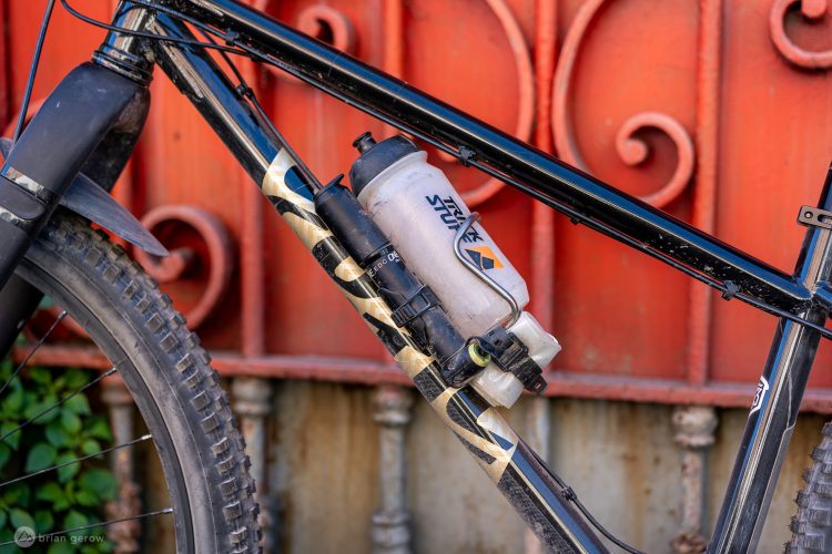 A close-up view of a mountain bike frame featuring a water bottle mounted on the triangle section. The bike has a black and patterned design, and the background shows a rustic red metal gate with decorative swirls. The bike's tire is partially visible, showcasing its tread pattern.