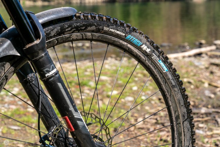 Close-up of a mountain bike's front wheel, featuring a partially muddy tire with visible tread patterns and branding. The fork and brake components are also visible, set against a blurred background of greenery and water.